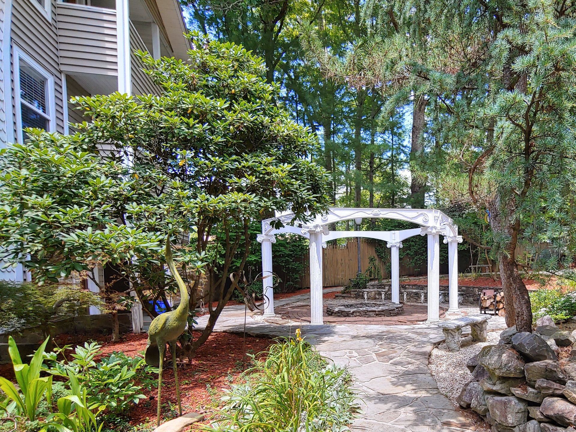 White garden arbor in a landscaped yard beside a house, surrounded by trees and shrubs.
