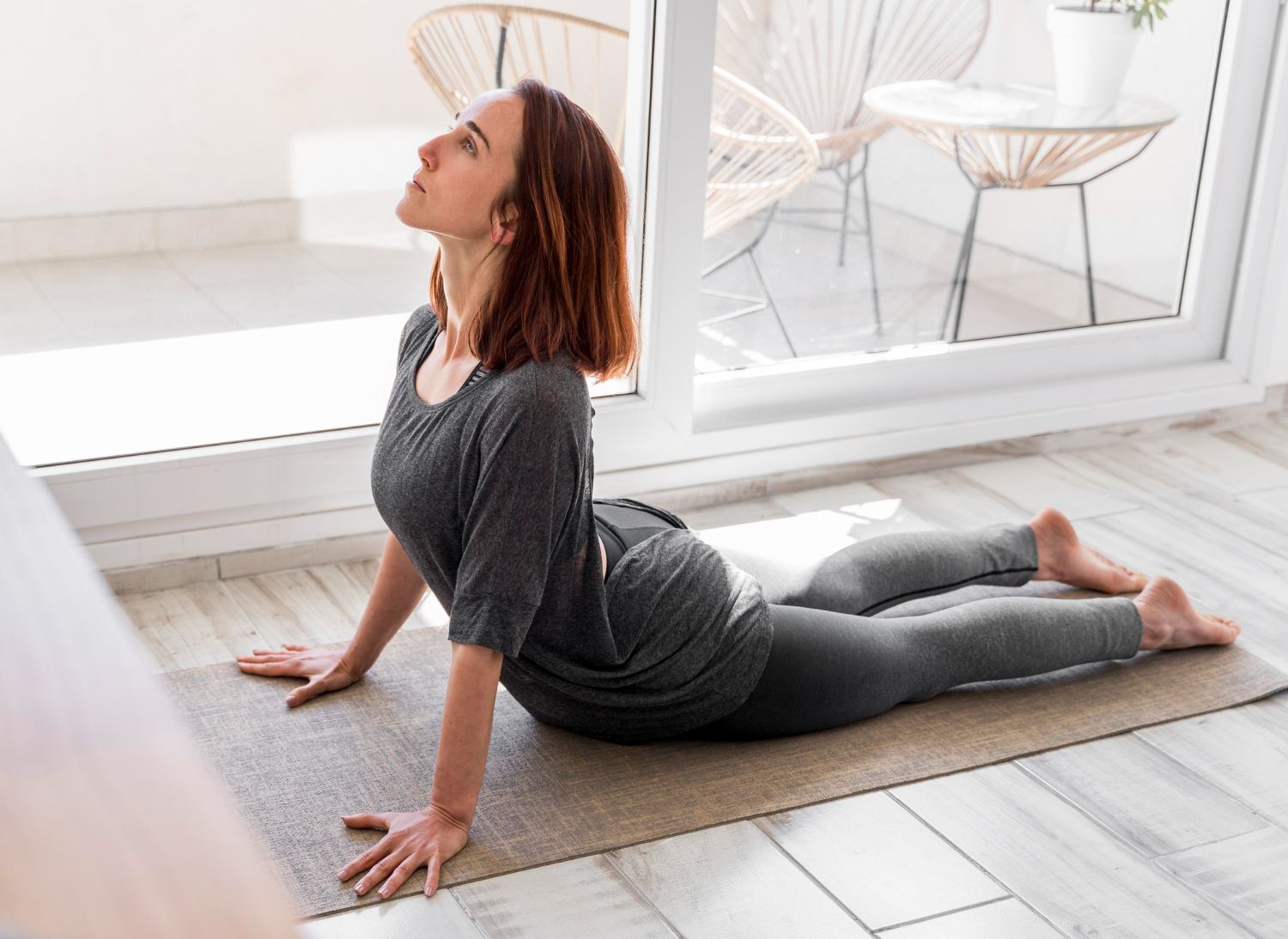 Woman doing upward-facing dog yoga pose on a mat in a bright room with white chairs and sliding doors