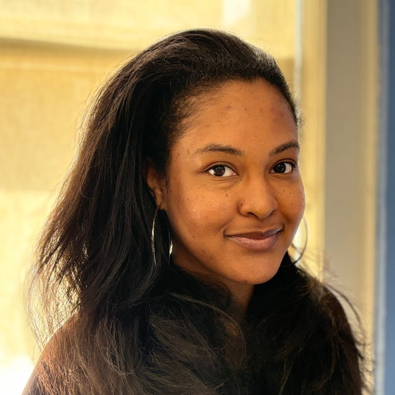 Smiling person with long dark hair standing indoors near a window in warm light
