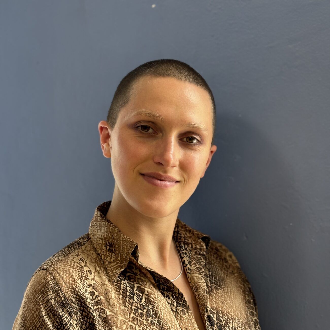 Smiling person with a shaved head in a patterned shirt against a blue wall