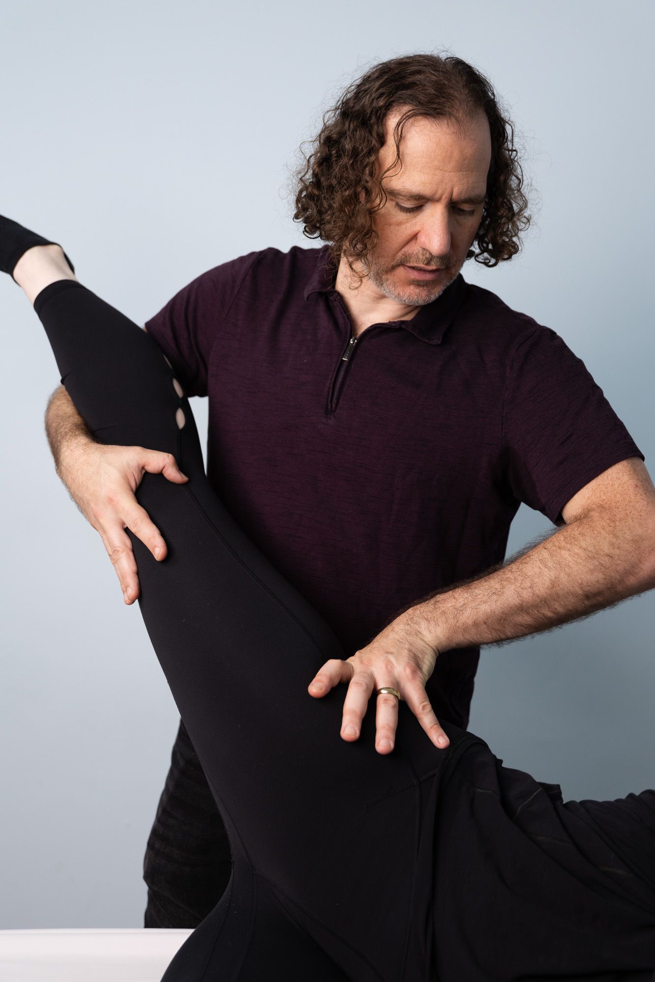 Man adjusting a dancer’s raised leg in a studio, both wearing dark clothing