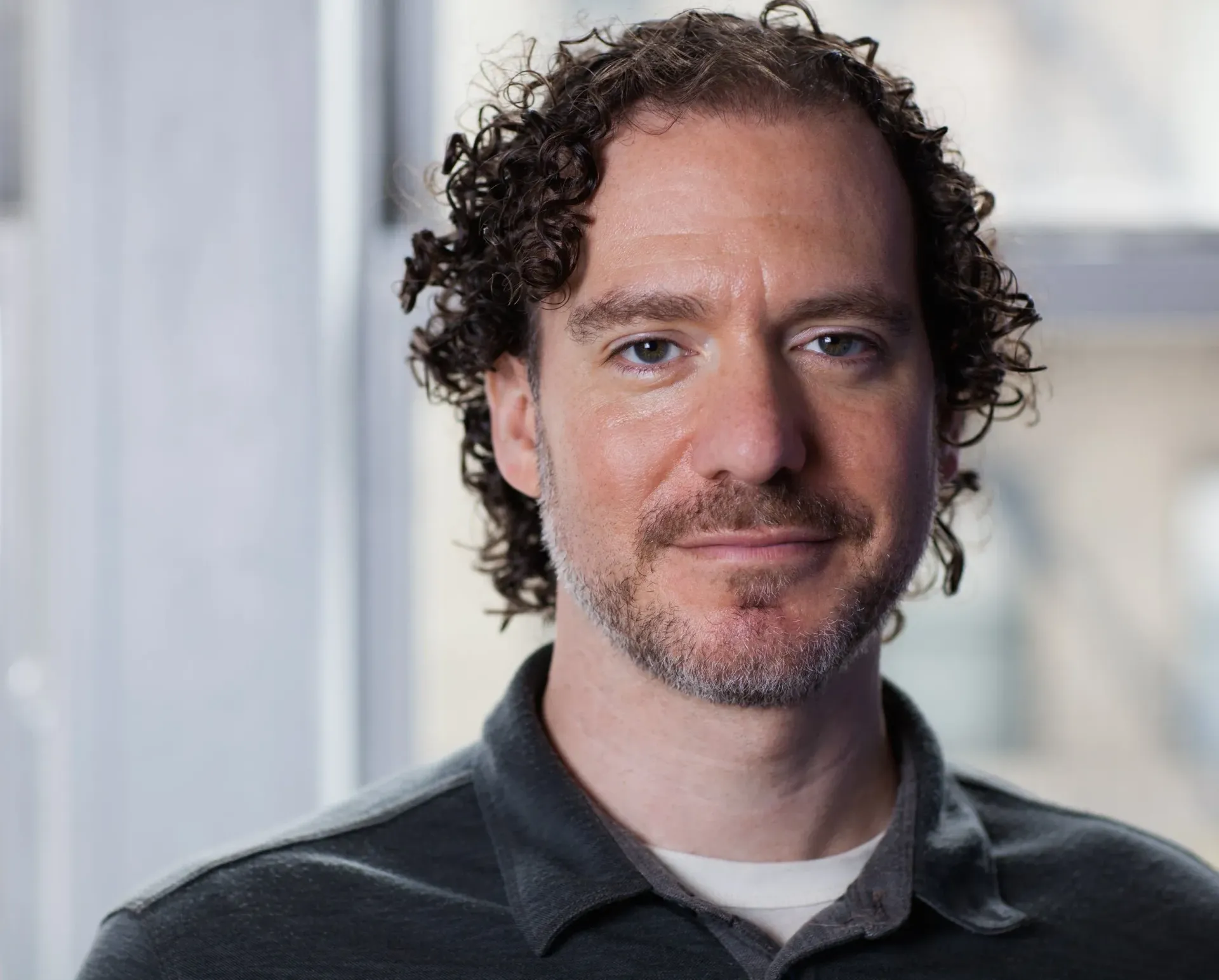 Portrait of a man with curly hair and a slight beard, wearing a dark shirt, against a blurred office background