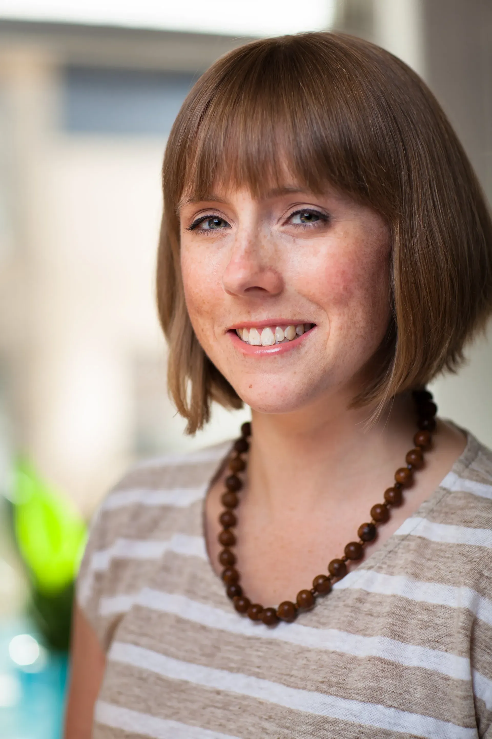 Woman with short brown hair and striped shirt smiling indoors, wearing a beaded necklace