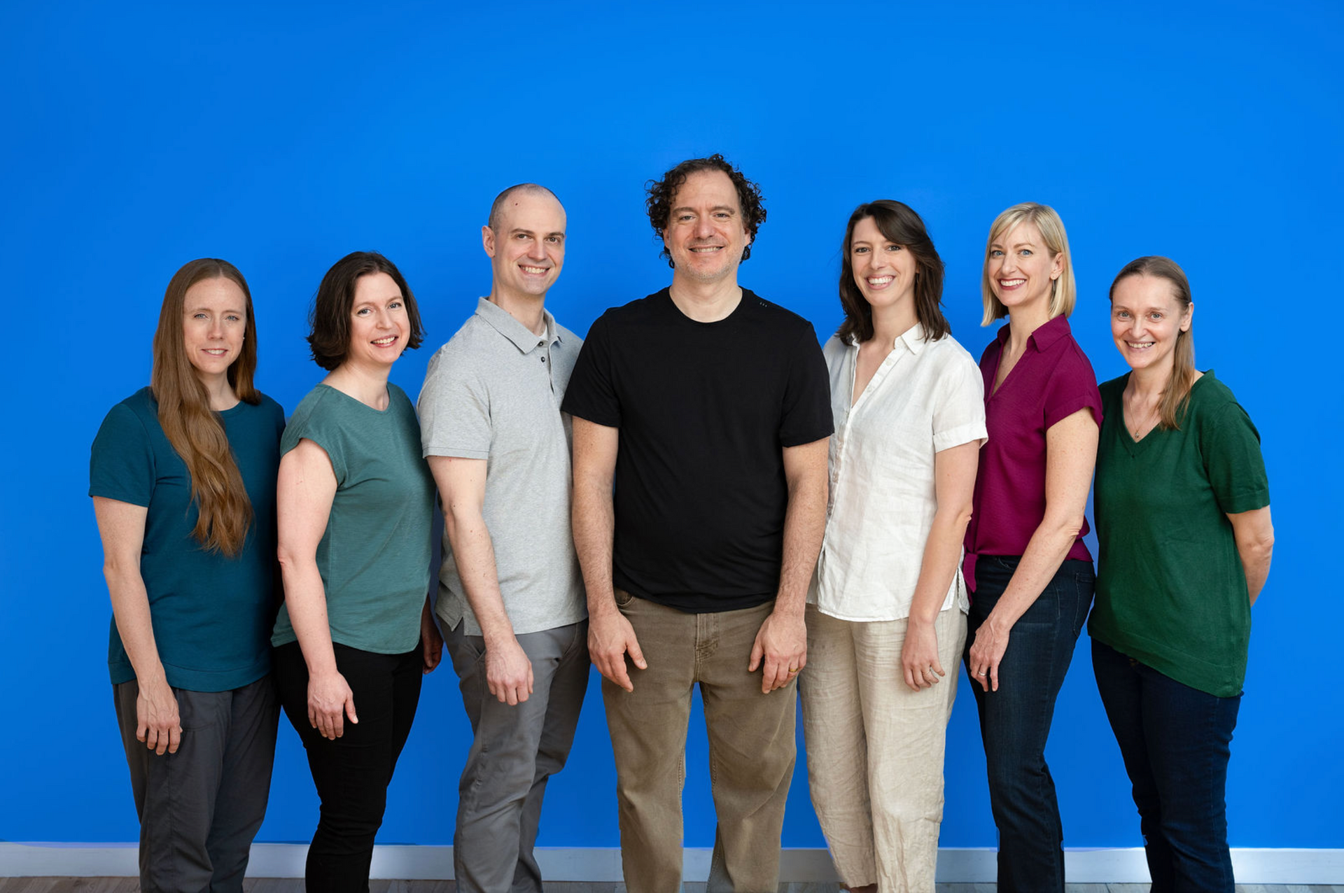 Seven people standing in a row against a bright blue background, smiling at the camera.