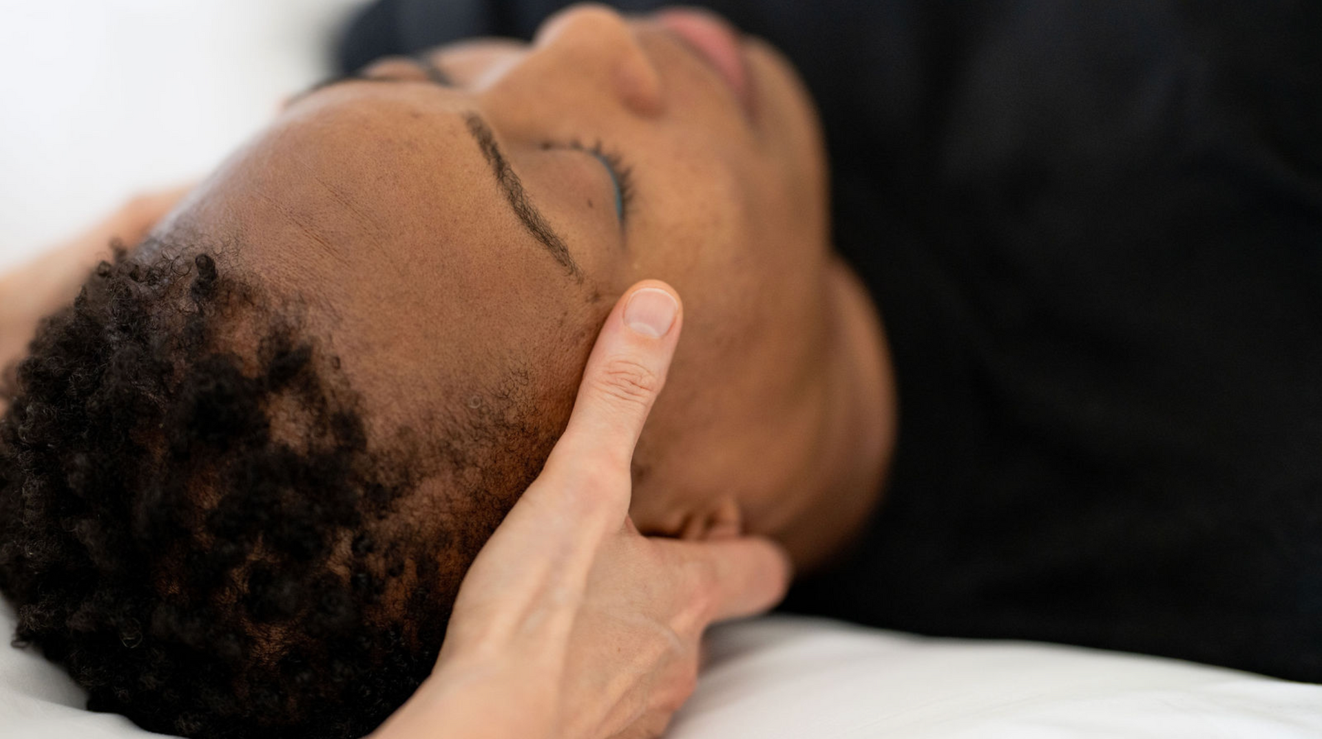 Hands gently massage a person's scalp on a cream treatment table in a bright room.