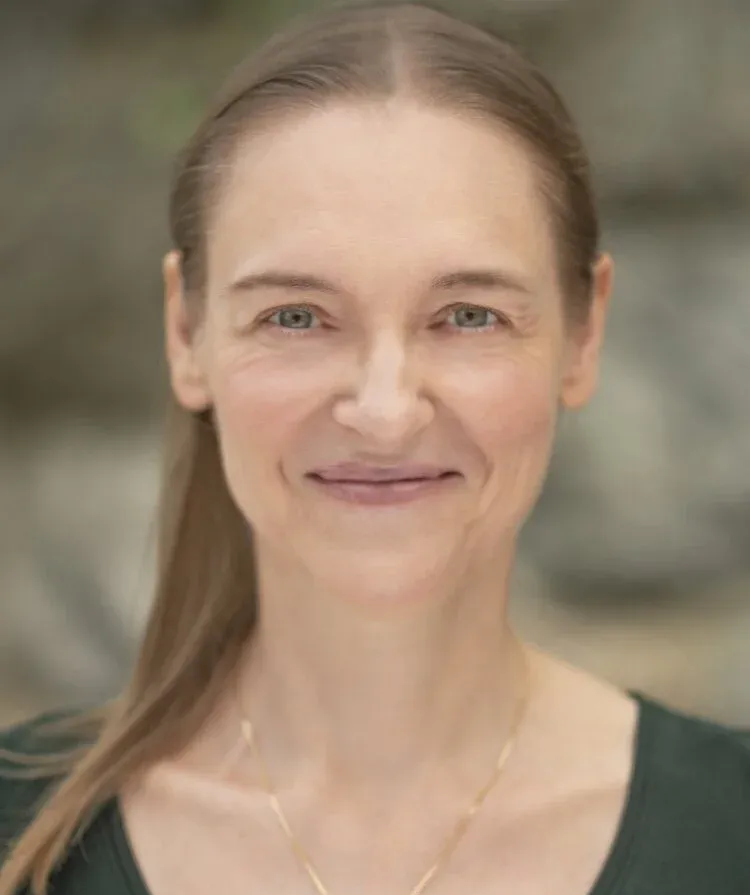 Smiling woman with light brown hair wearing a dark green top and a gold necklace against a neutral background