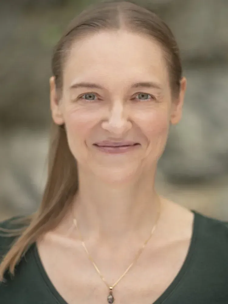Portrait of a smiling person wearing a dark green top and gold necklace against a soft background