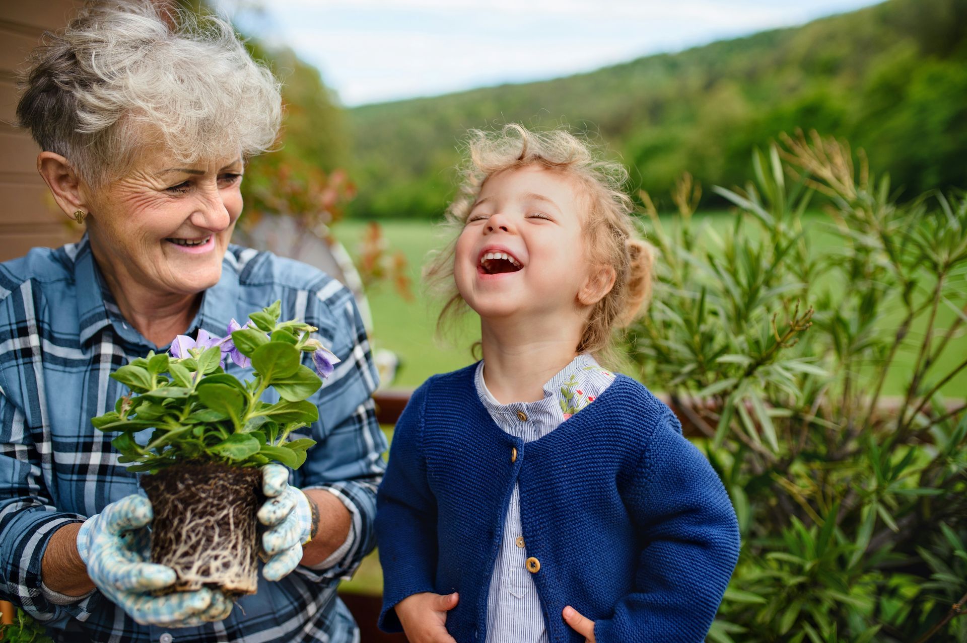 Two people smiling in a garden, holding a potted plant and laughing together.