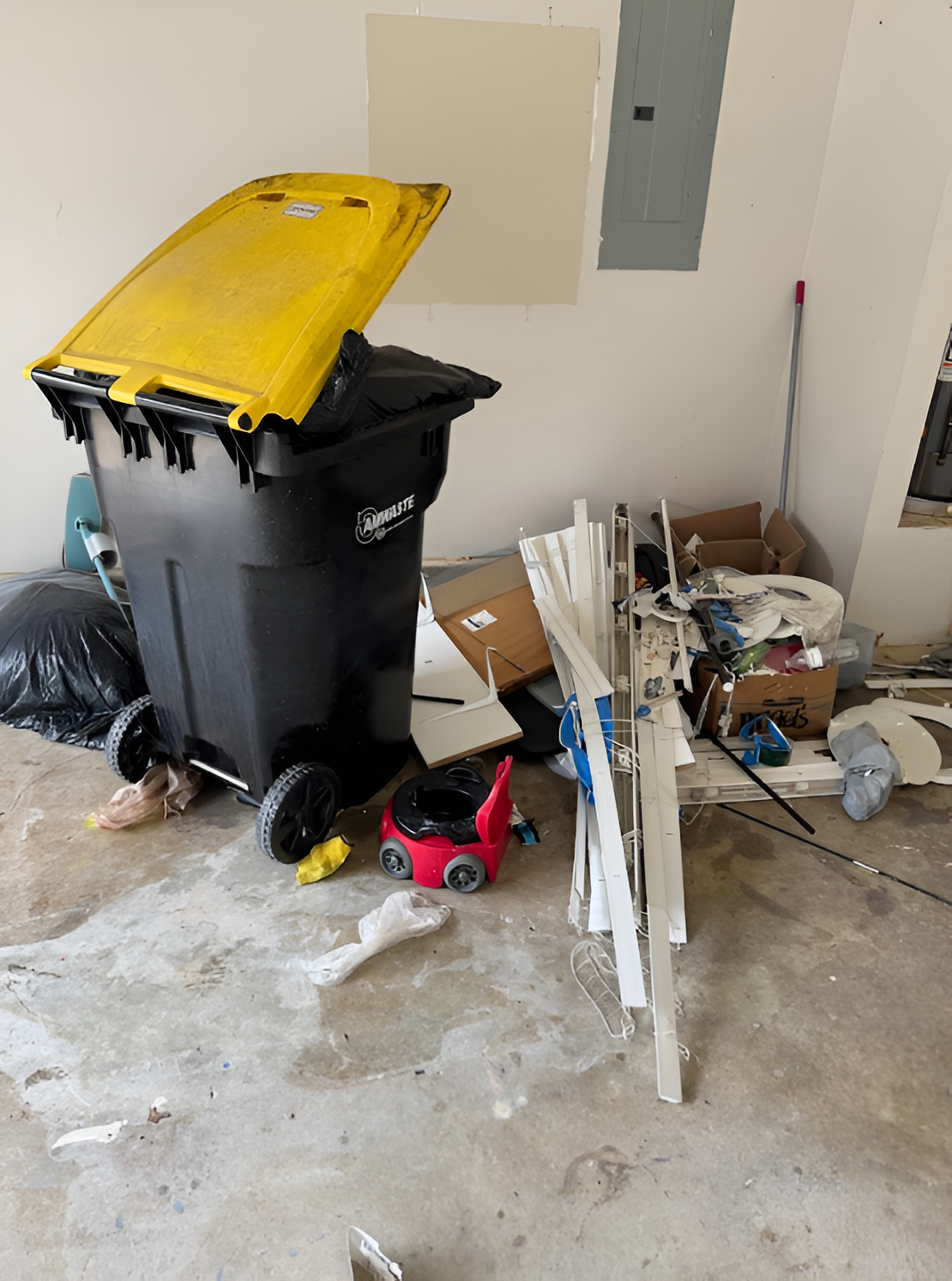 Cluttered garage interior with trash bin, debris, and tools on a concrete floor, near a partially painted wall.