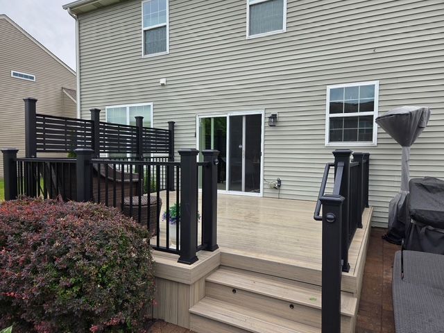 Backyard deck with steps, sliding glass door, black railings, and a covered grill beside a shrub.