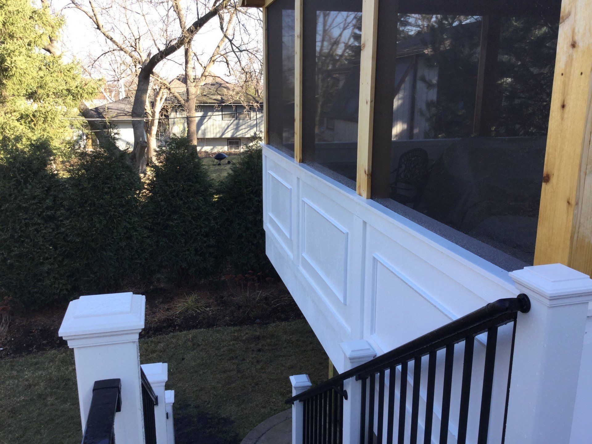 Screened porch with white railing and black stairs beside a hedge and lawn.