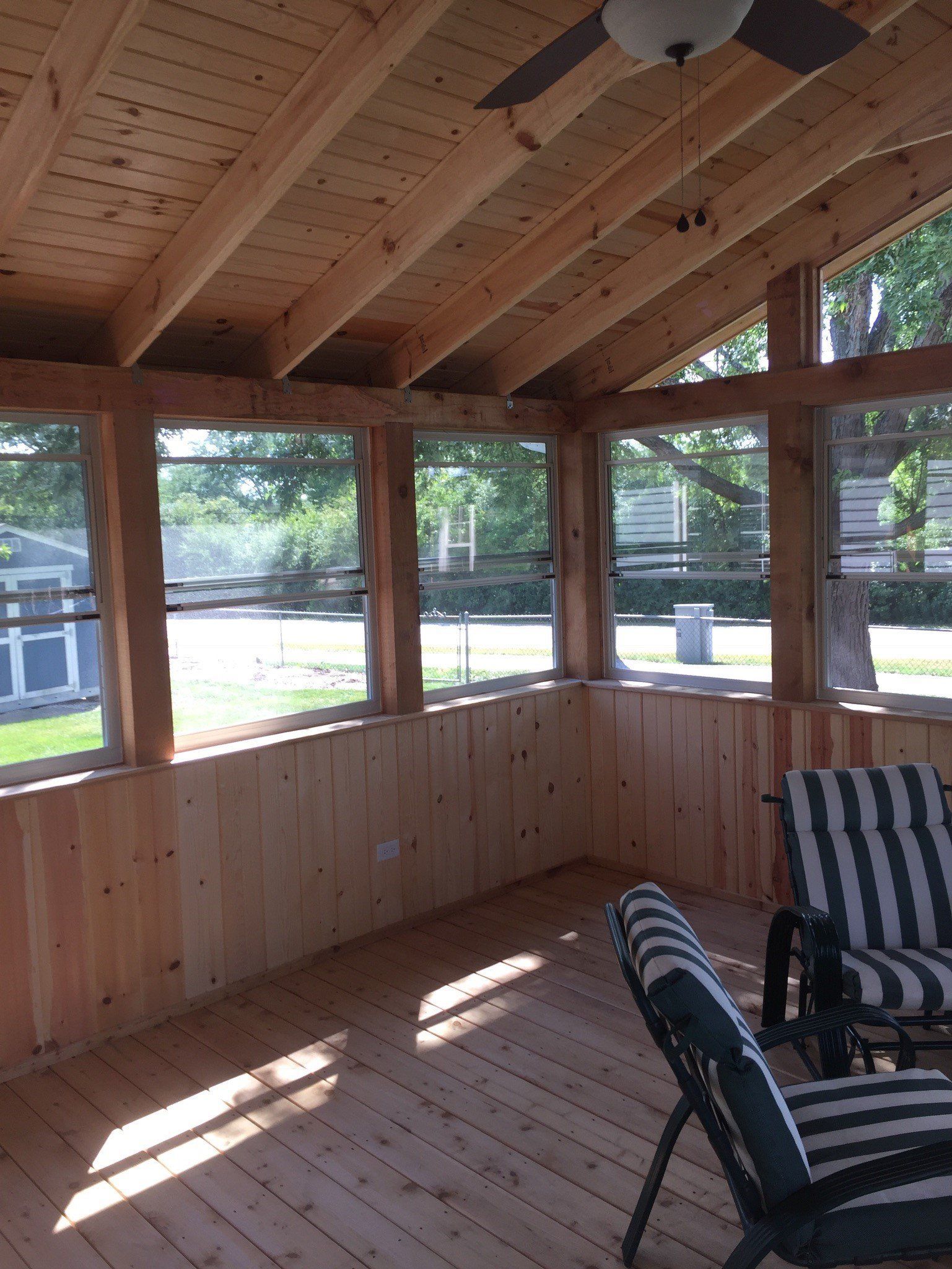 Sunlit screened porch with wooden walls, ceiling fan, and striped chairs overlooking a yard