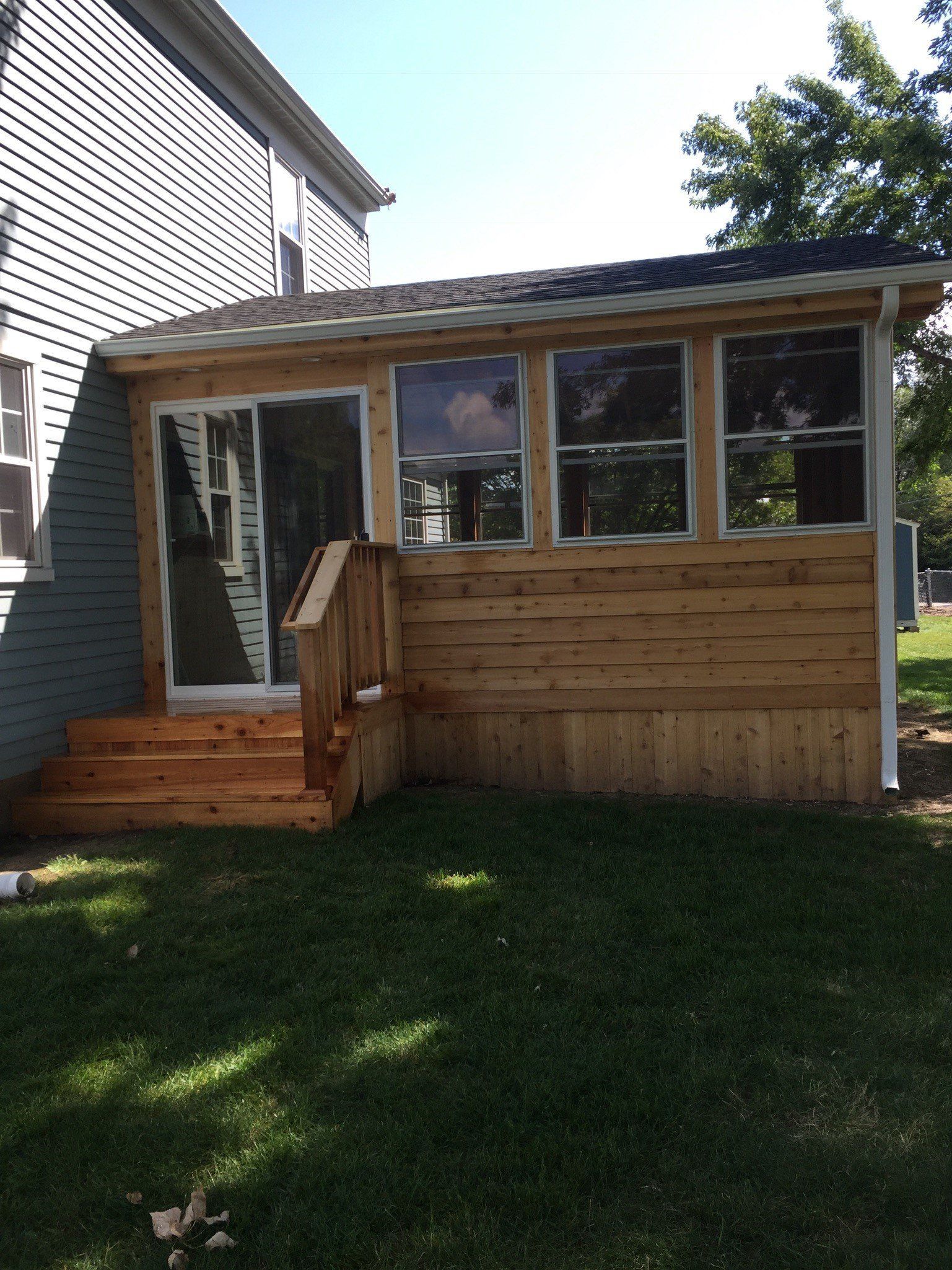 Backyard wooden sunroom with windows and a small porch beside a house, overlooking a grassy lawn