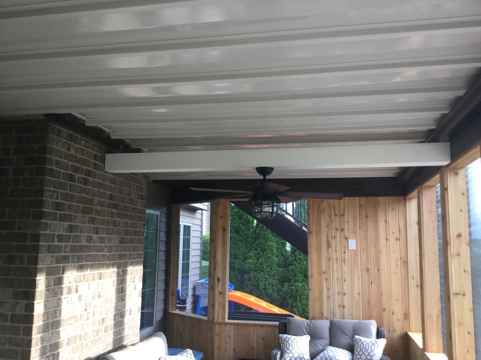 Covered patio with gray sofa, wooden fence, brick wall, and ceiling fan under a corrugated roof.