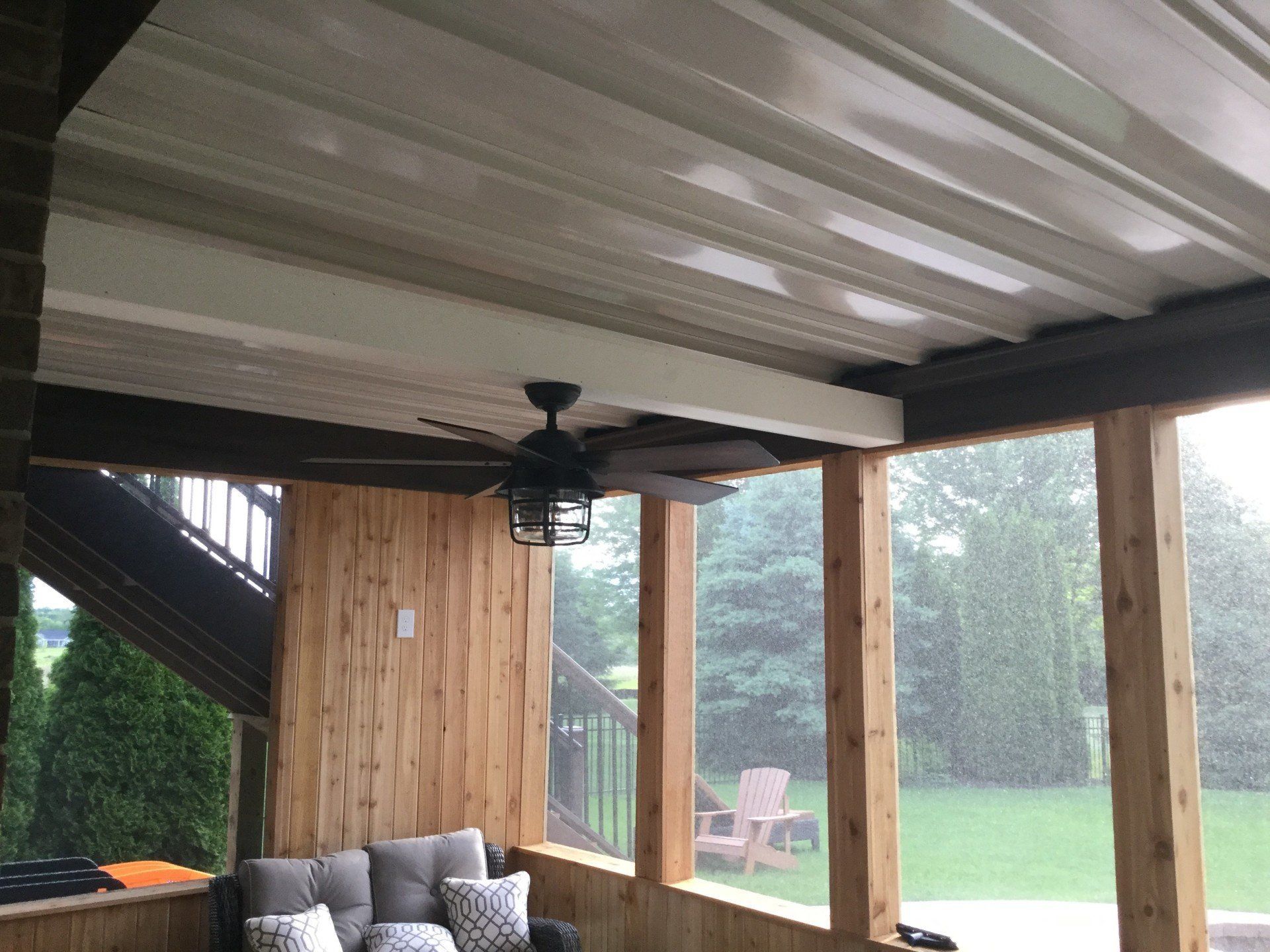 Covered patio with wood posts, ceiling fan, couch, and a view of a green yard in the rain