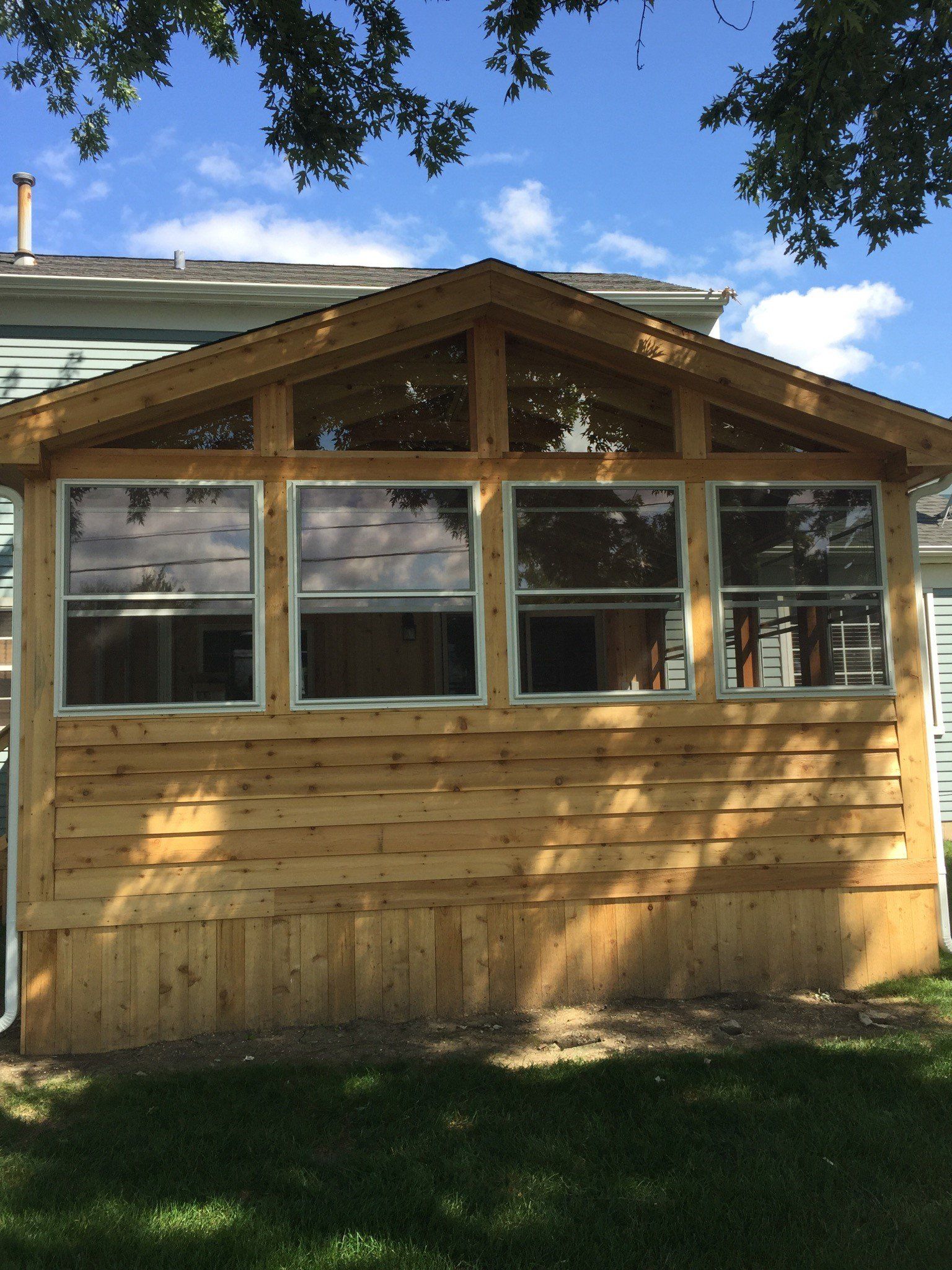 Small wooden sunroom with windows and a peaked roof, set in a grassy yard under a blue sky.