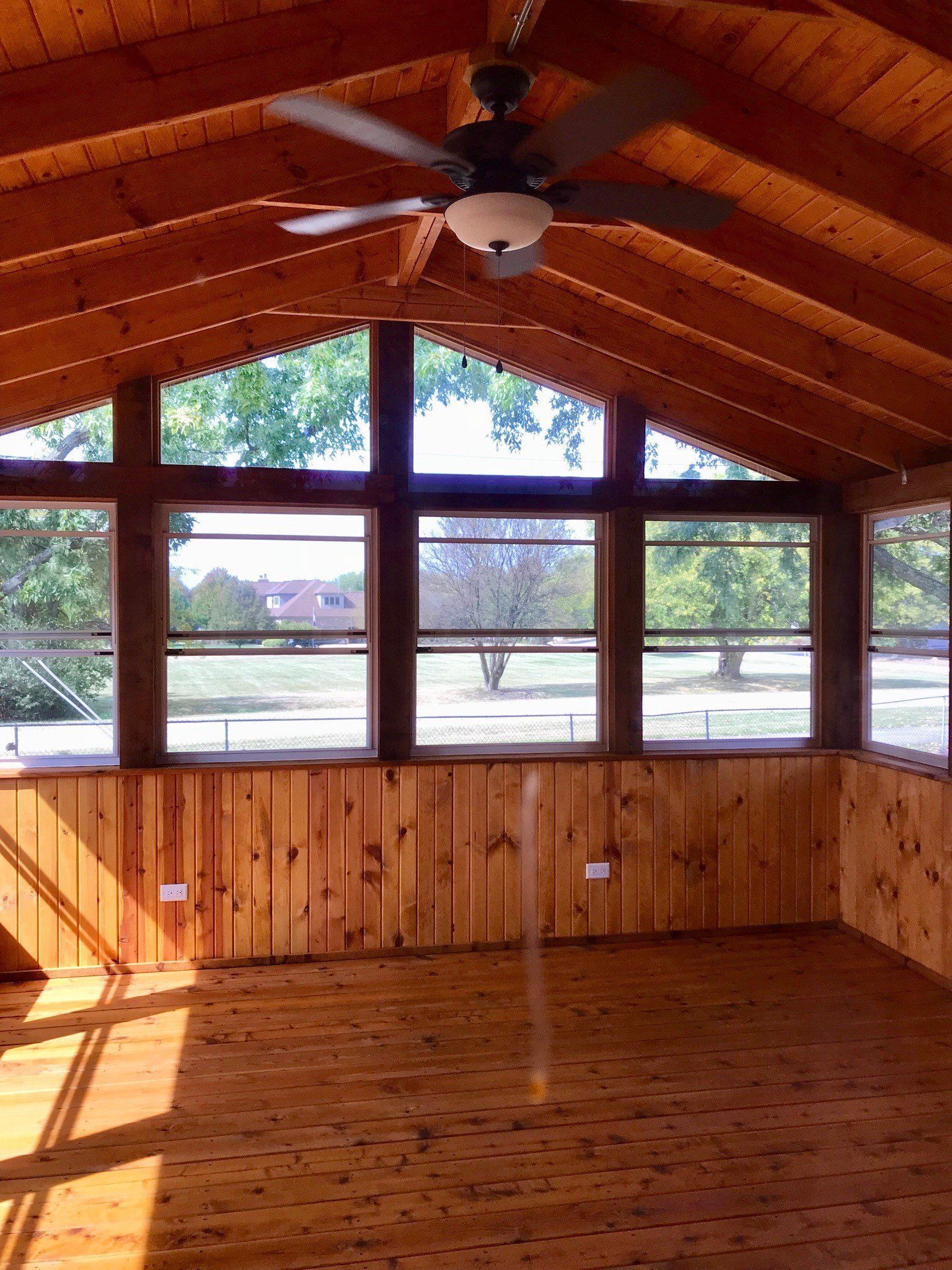 Wood-paneled sunroom with large windows, vaulted cedar ceiling, and a ceiling fan.