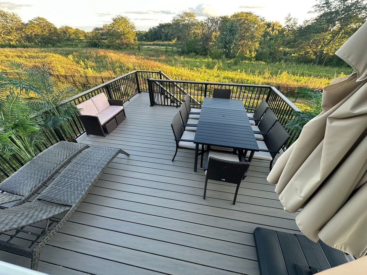 Outdoor deck with black patio table and chairs, cushioned bench, and a view of green fields at sunset