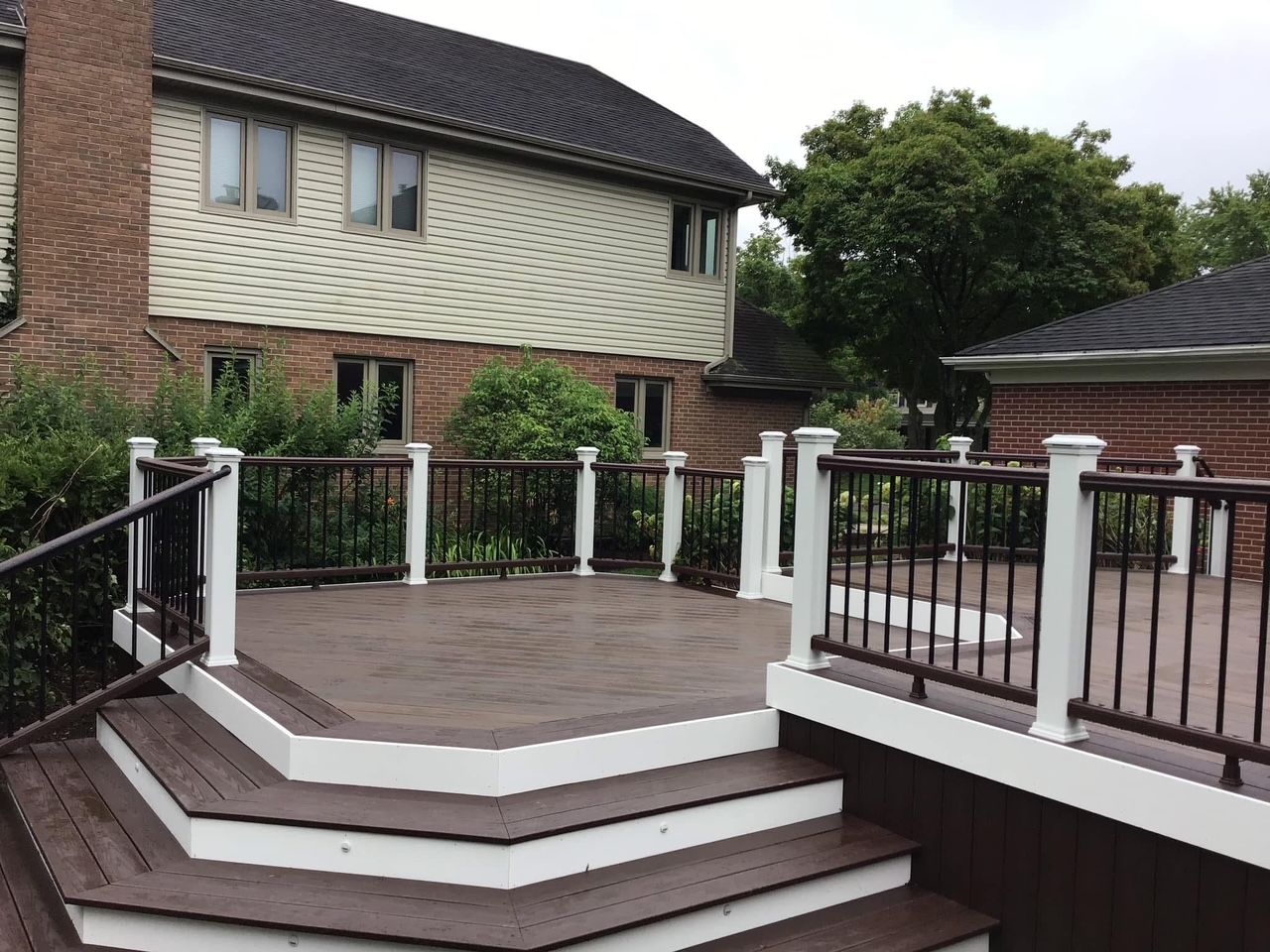 Raised patio deck with white railings and steps beside a brick house and trees
