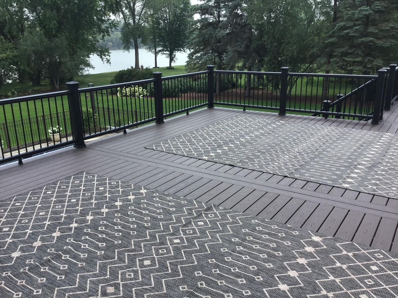 Outdoor viewing deck with black railings, patterned pavement, and trees by a lake.