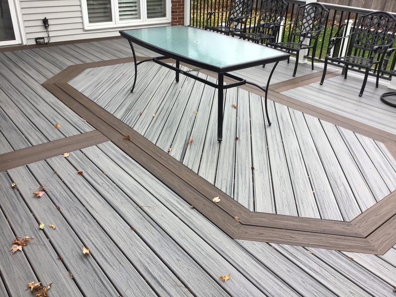 Wooden deck with a glass-top table and black chairs beside a house.