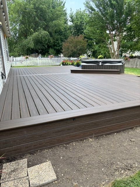Brown wooden deck with a raised hot tub, bordered by a white fence and green trees