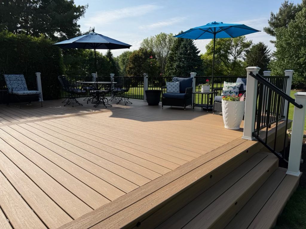 Sunlit wooden deck with patio tables, blue umbrellas, and potted plants overlooking a garden