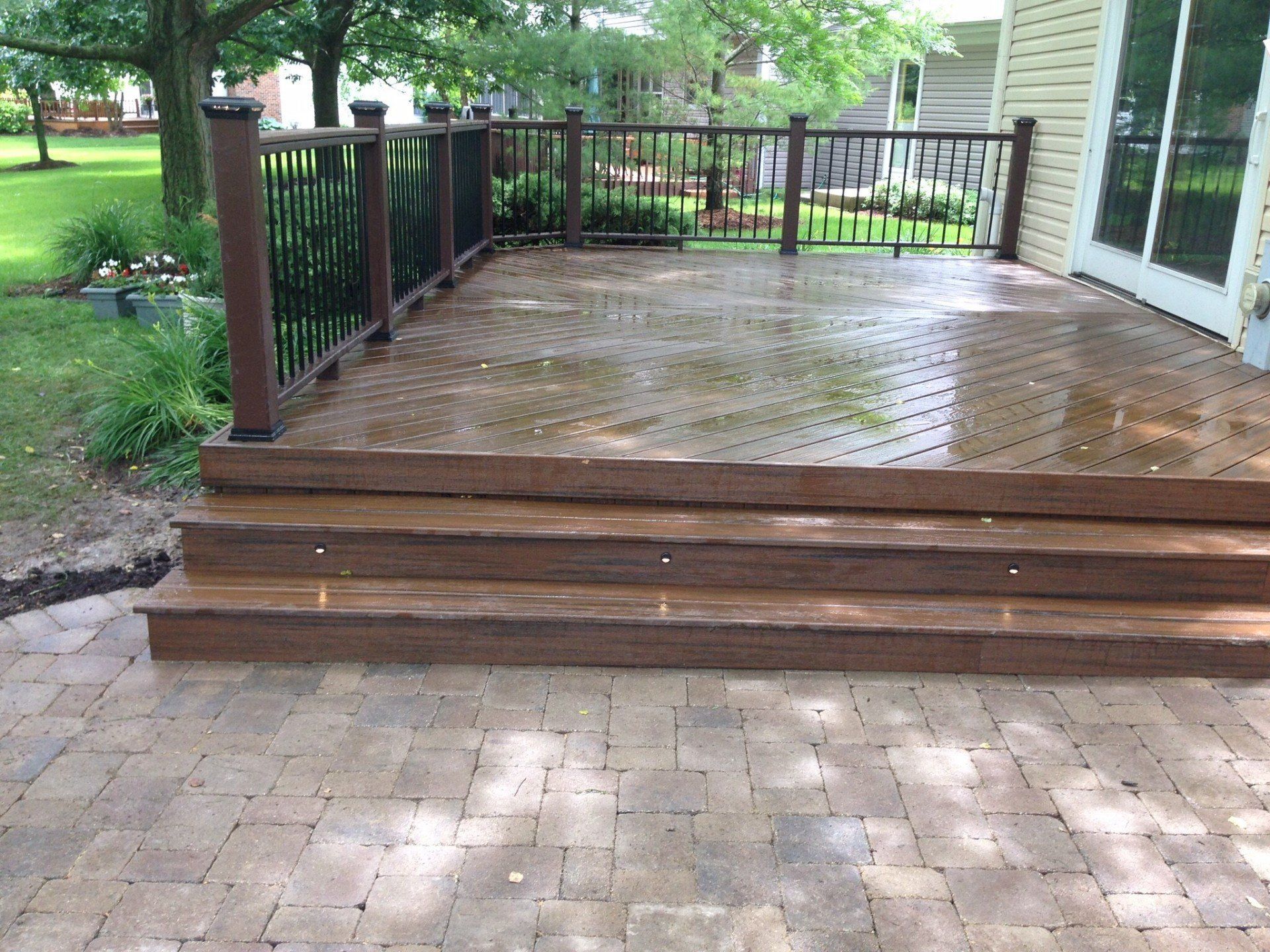 Wooden deck with wet boards, black railing, and three steps leading down to a brick patio.