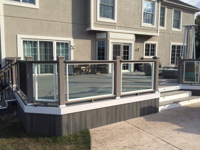 Raised glass-railed deck attached to a beige house, with steps leading to a concrete patio.