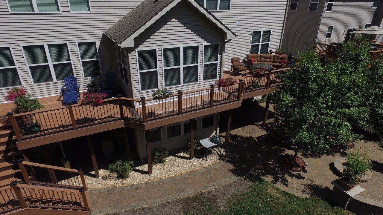 Back deck with railing and chairs beside a house, overlooking a landscaped yard and tree.