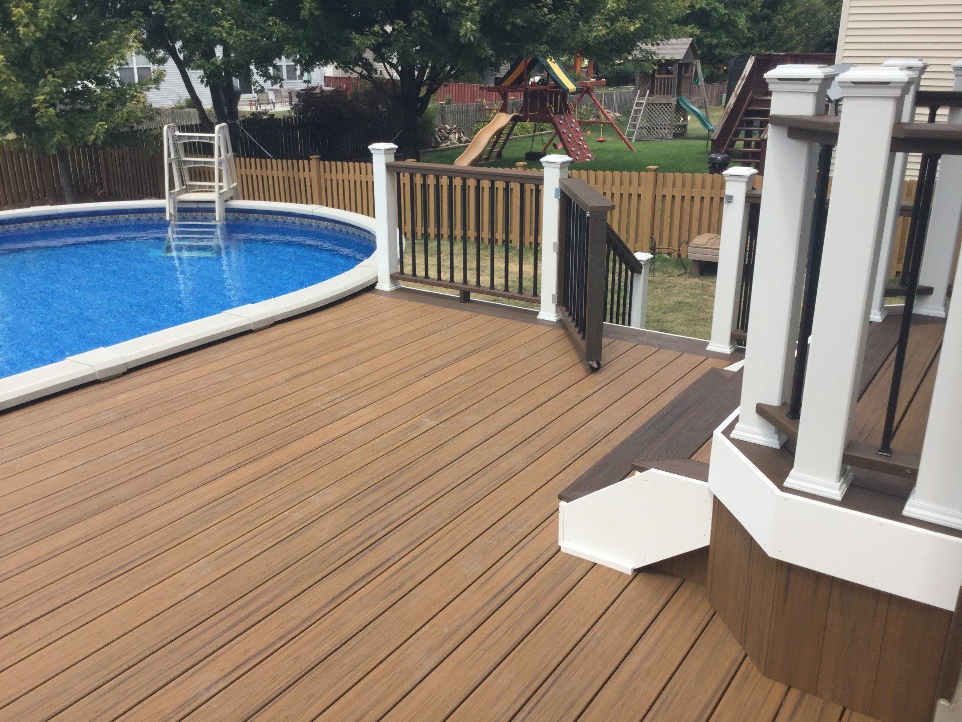 Wooden pool deck with white railing beside a blue swimming pool and a backyard fence.