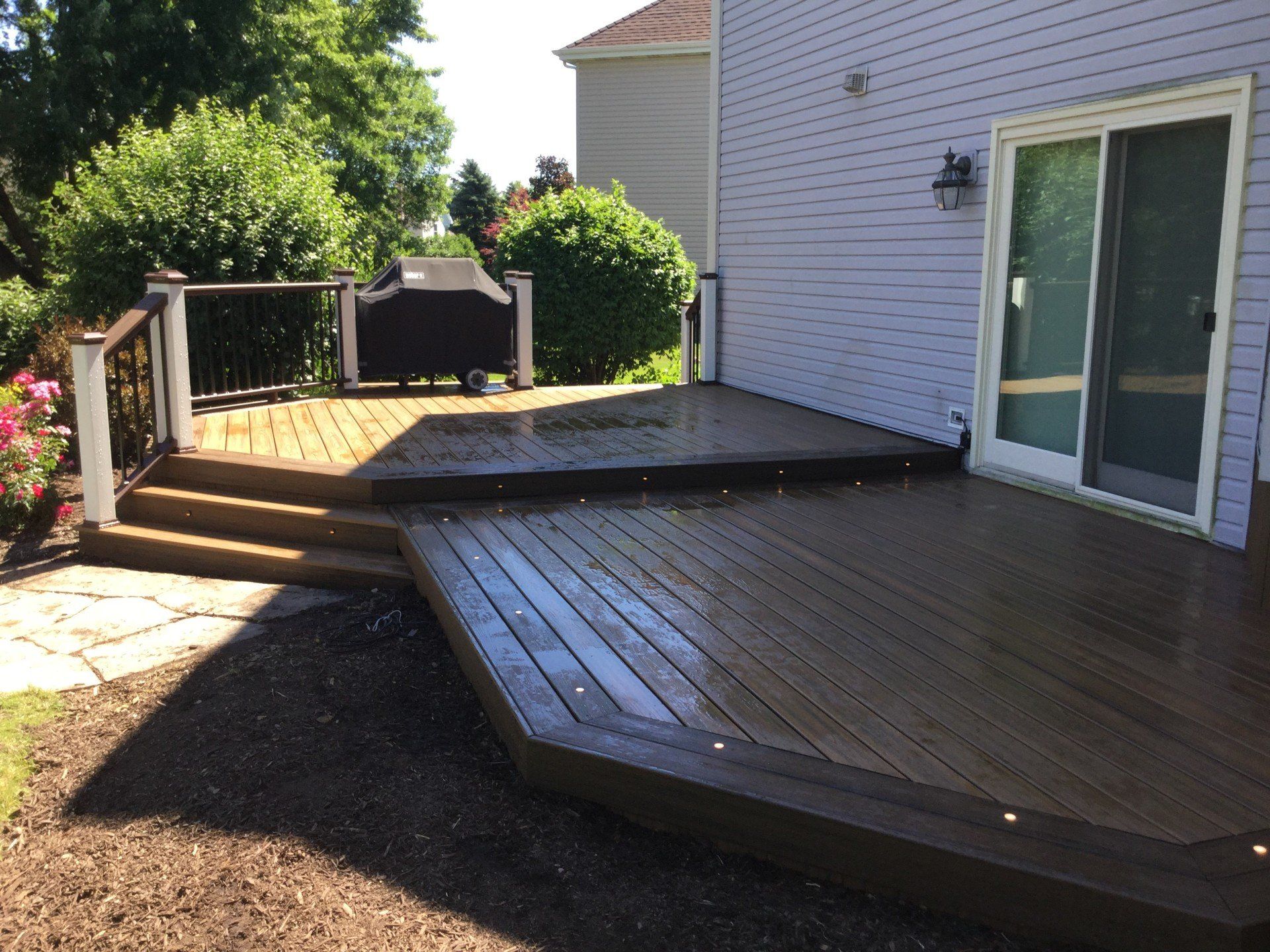 Wooden backyard deck beside a house, with steps, railing, and sliding glass doors.