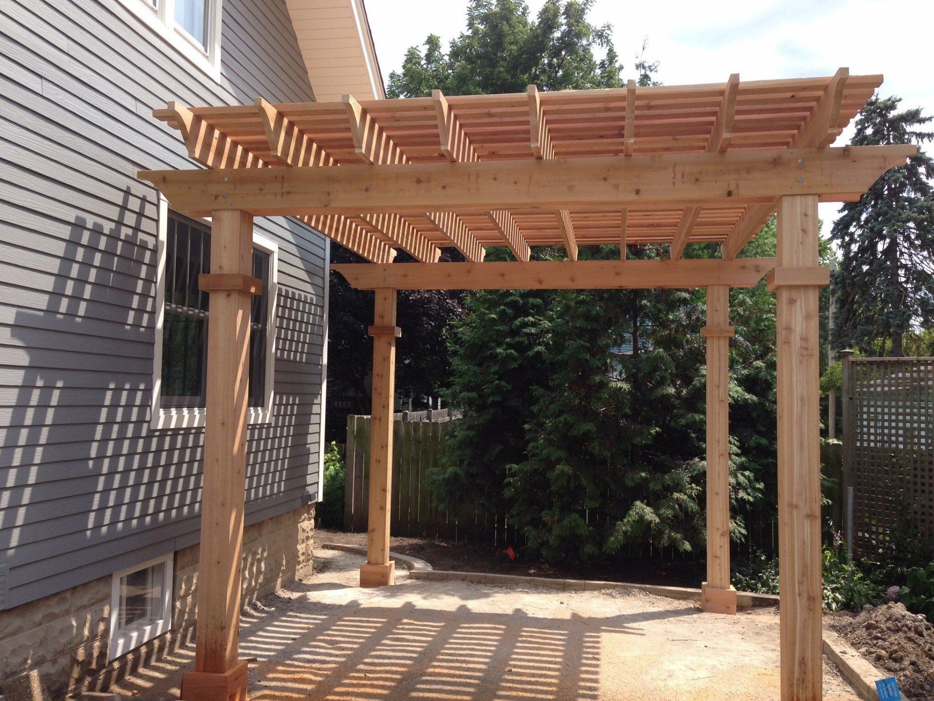 Wooden pergola-covered patio beside a house, with slatted roof and concrete floor.