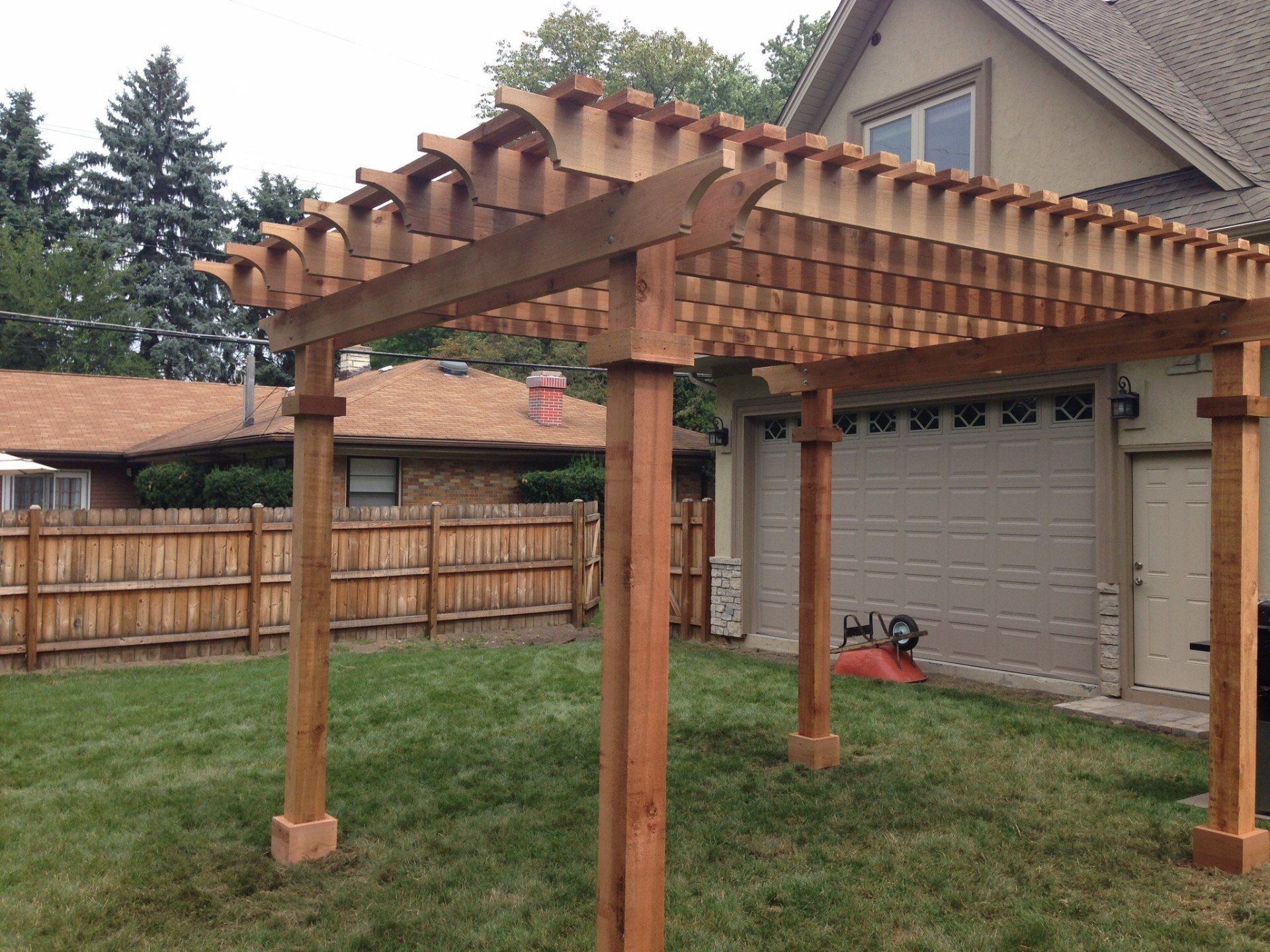 Wooden pergola in a backyard beside a garage, with grass yard and fenced perimeter
