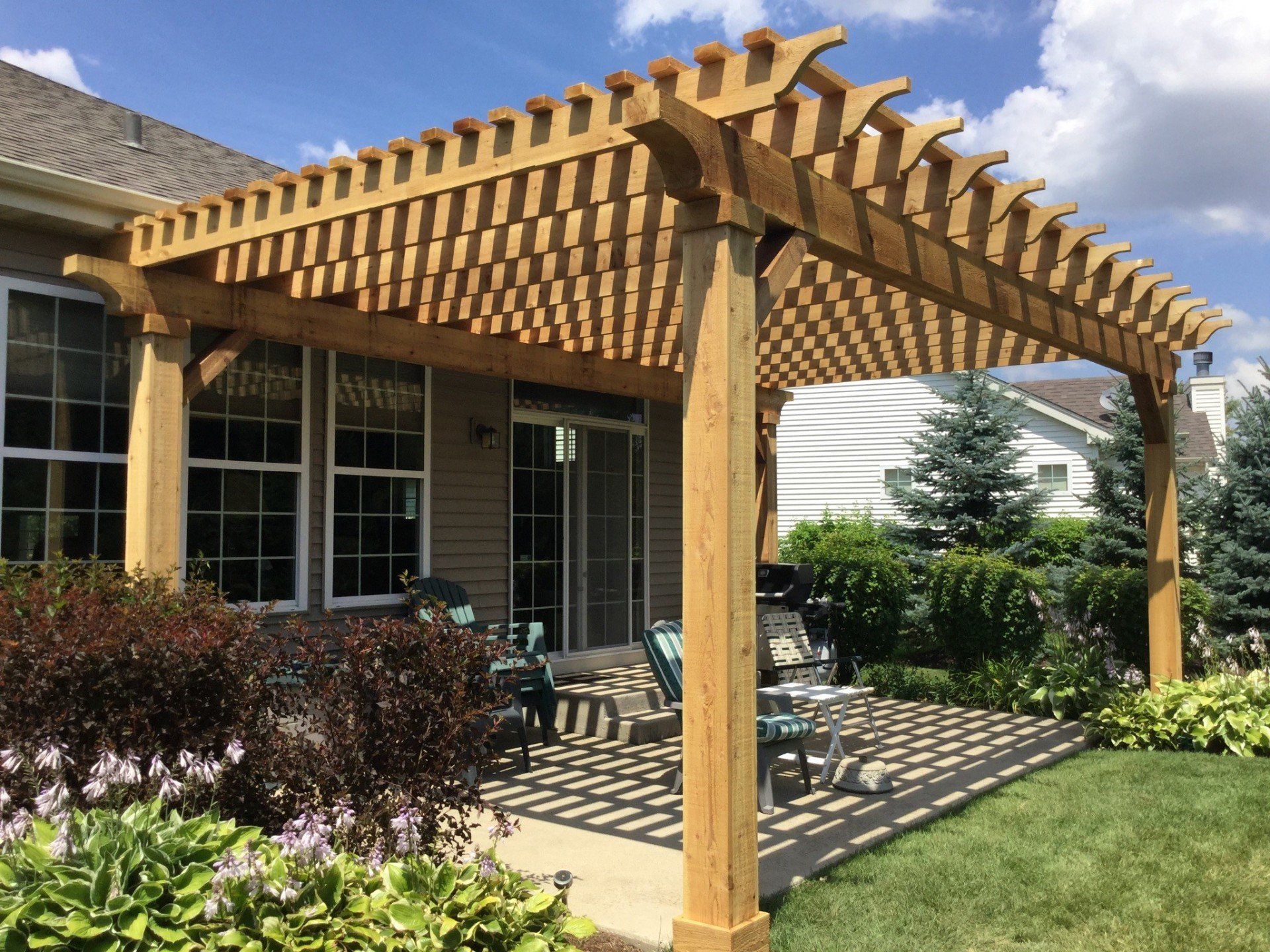 Wooden pergola over a patio beside a brick house, with sunlight casting lattice shadows on the grass.