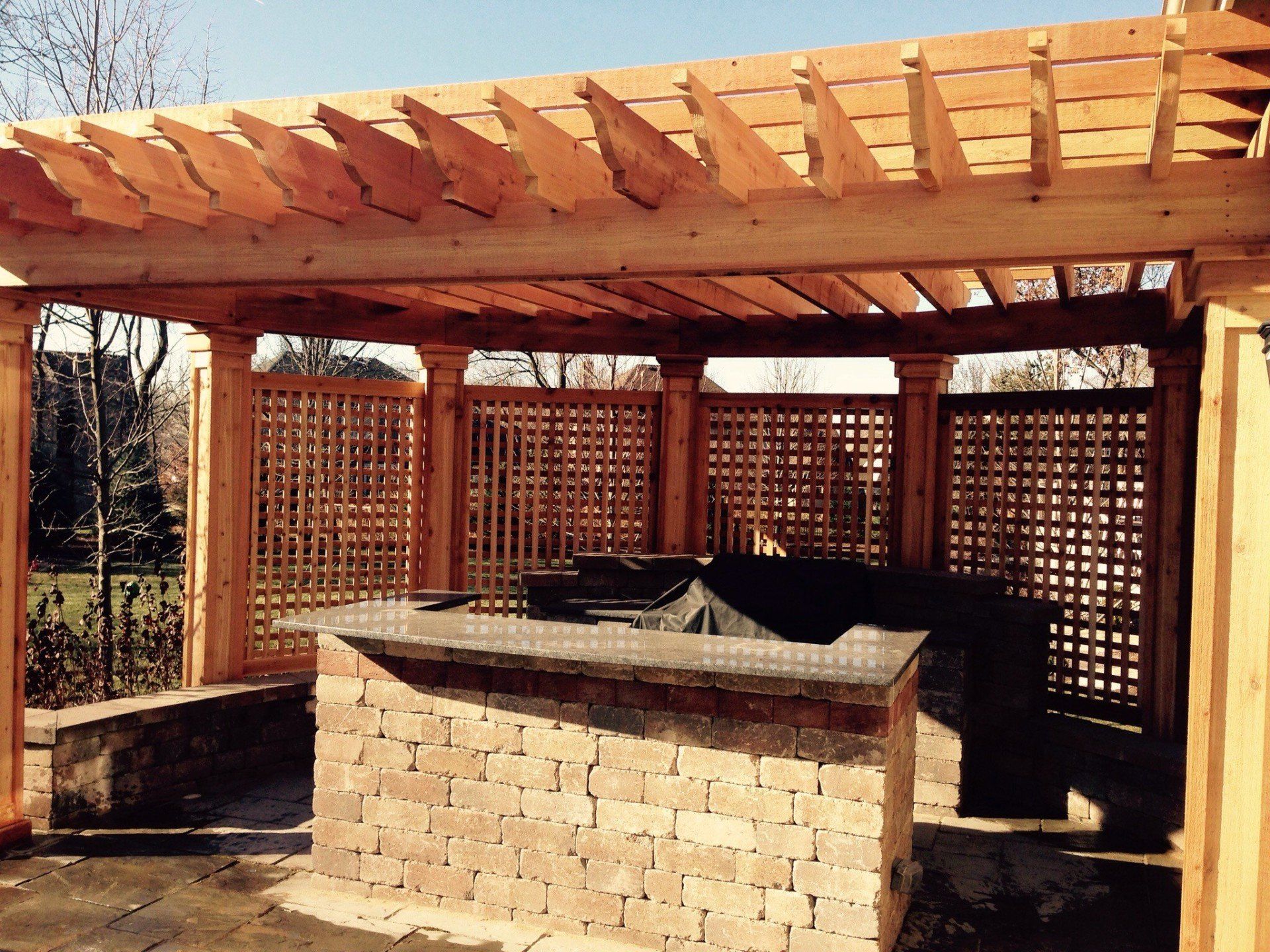 Brick outdoor grill under a wooden pergola with lattice side panels in a backyard.