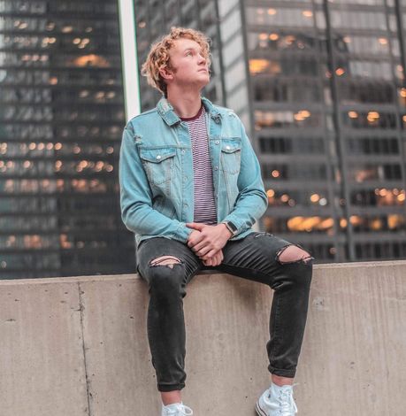 A young man in a denim jacket is sitting on a wall in front of a city skyline.