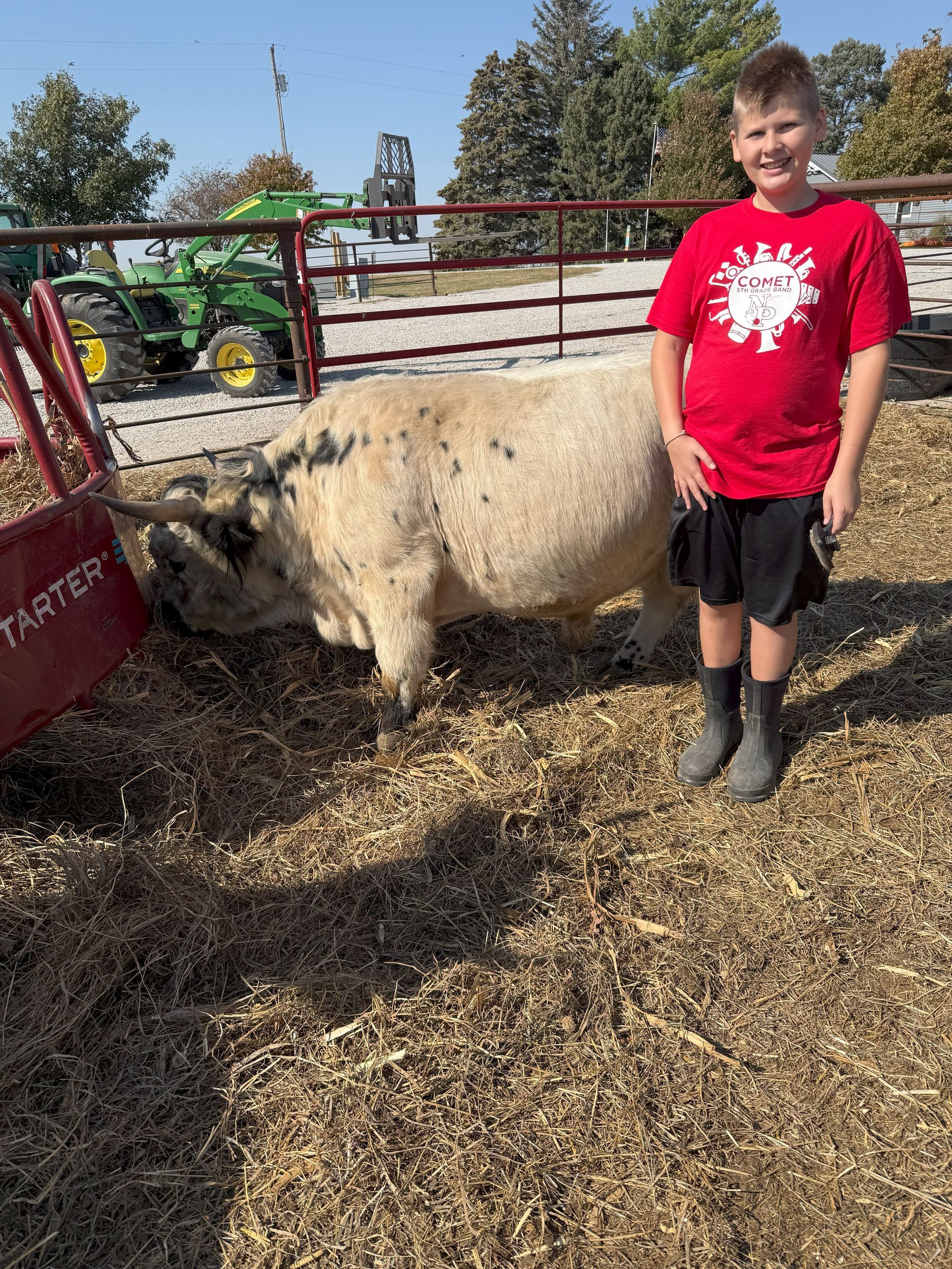 Boy in red shirt stands beside a white goat in a farm pen with hay and a green tractor in the background.