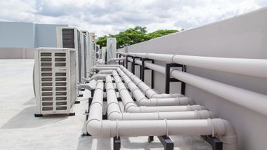 Rooftop HVAC pipes and air conditioners lined along a white wall under a cloudy sky