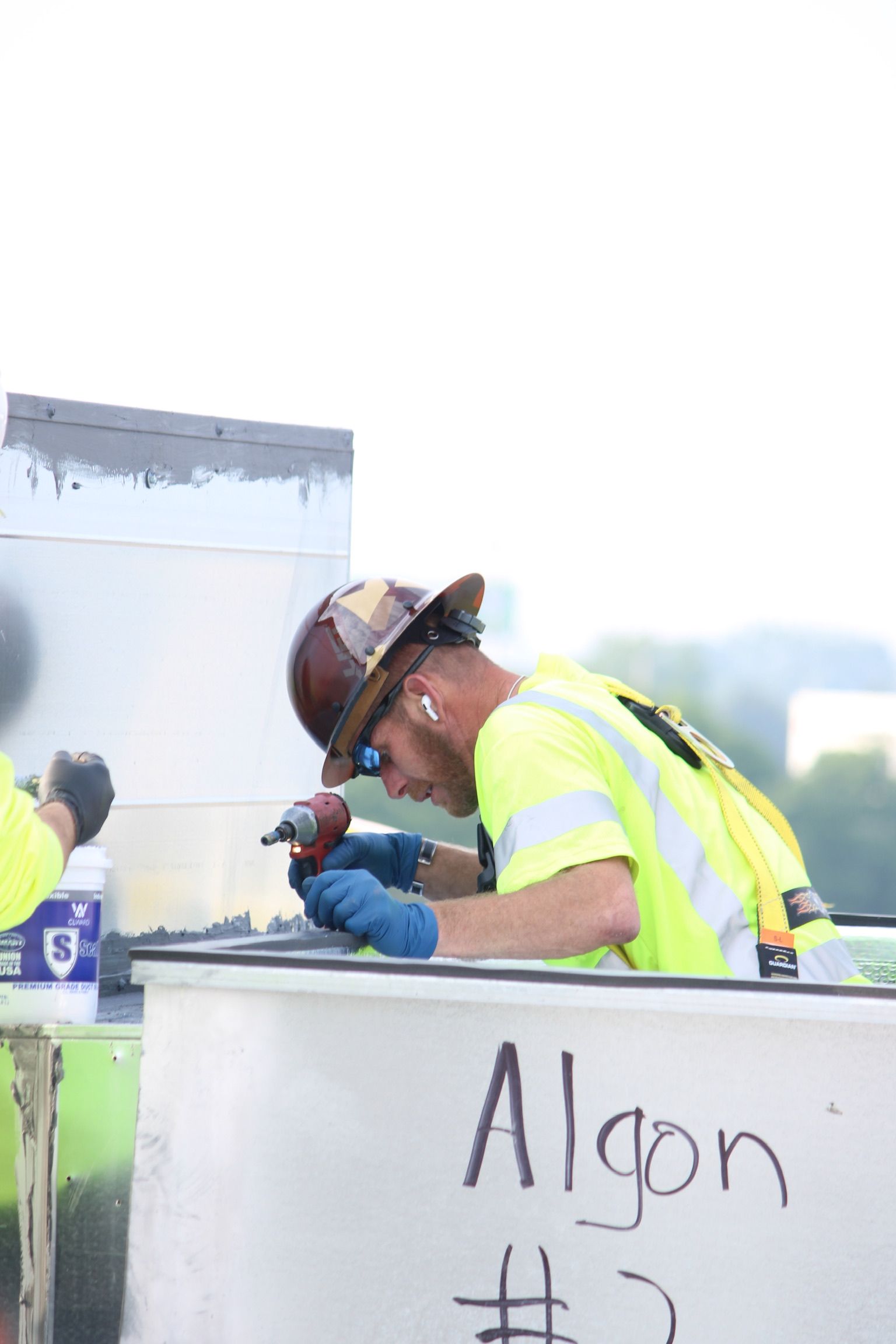 Worker in neon safety vest and hard hat crouches over a container, writing on it with a marker.