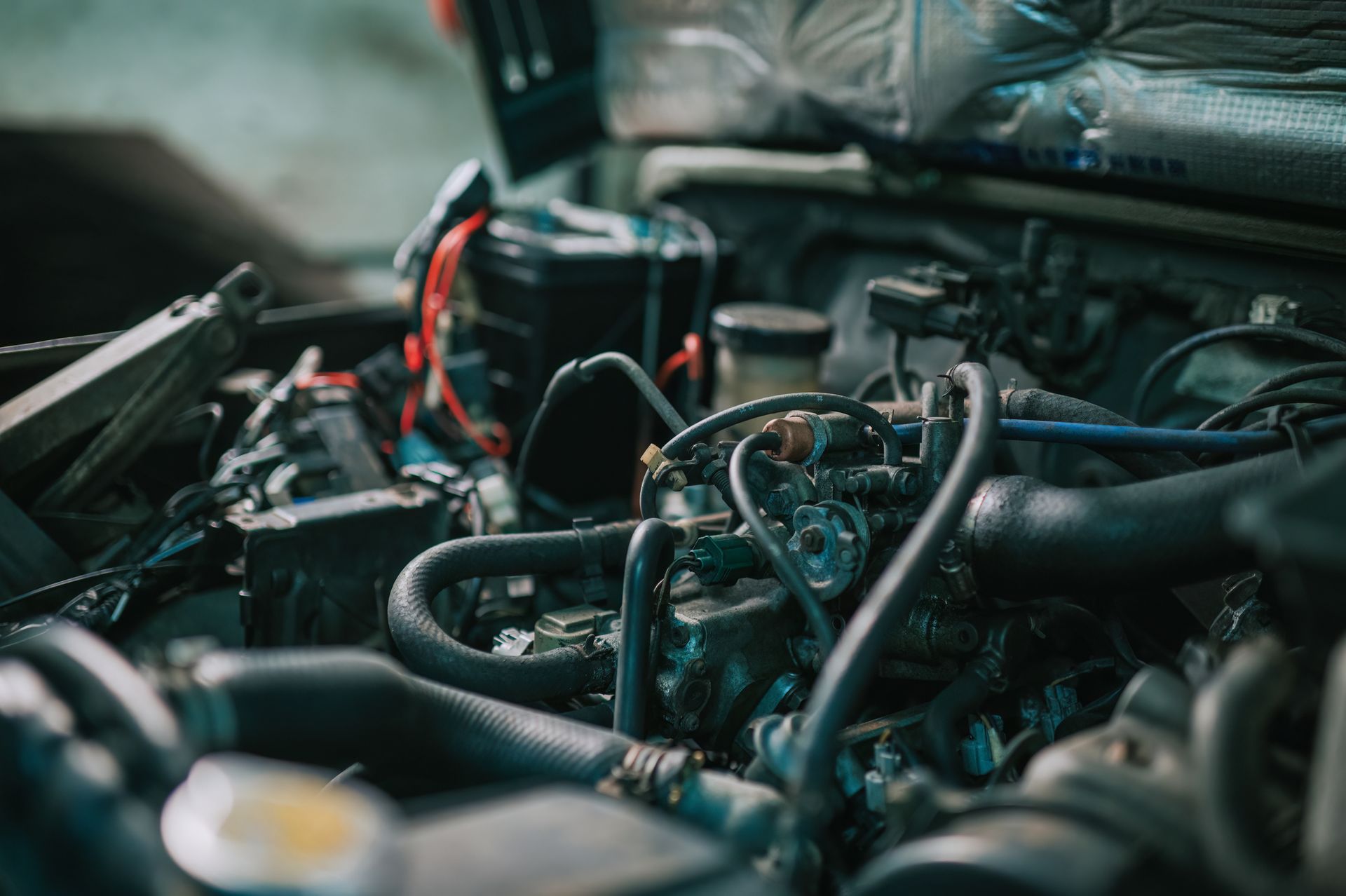 Car engine with hoses, wires, and battery in a workshop setting.