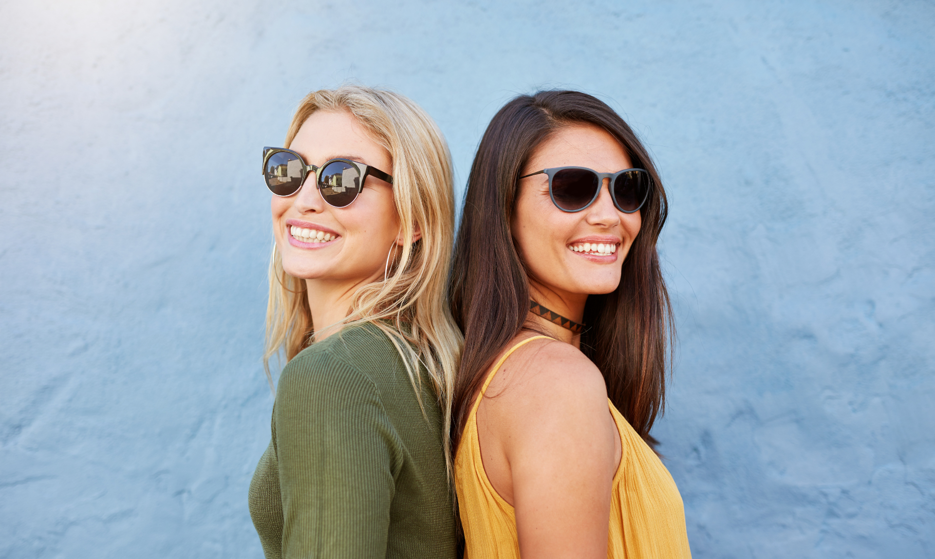 Dos mujeres que llevan gafas de sol están paradas espalda con espalda frente a una pared azul.