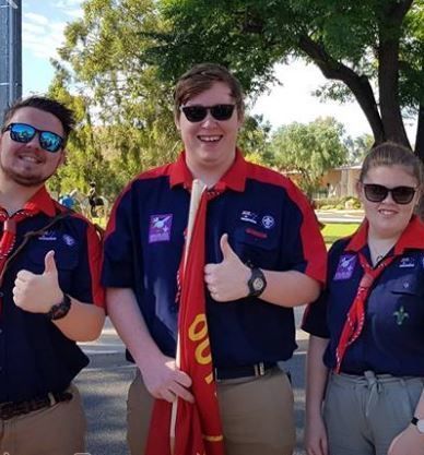 Three people in scout uniforms give thumbs up outdoors, holding a red flag, sunny day.