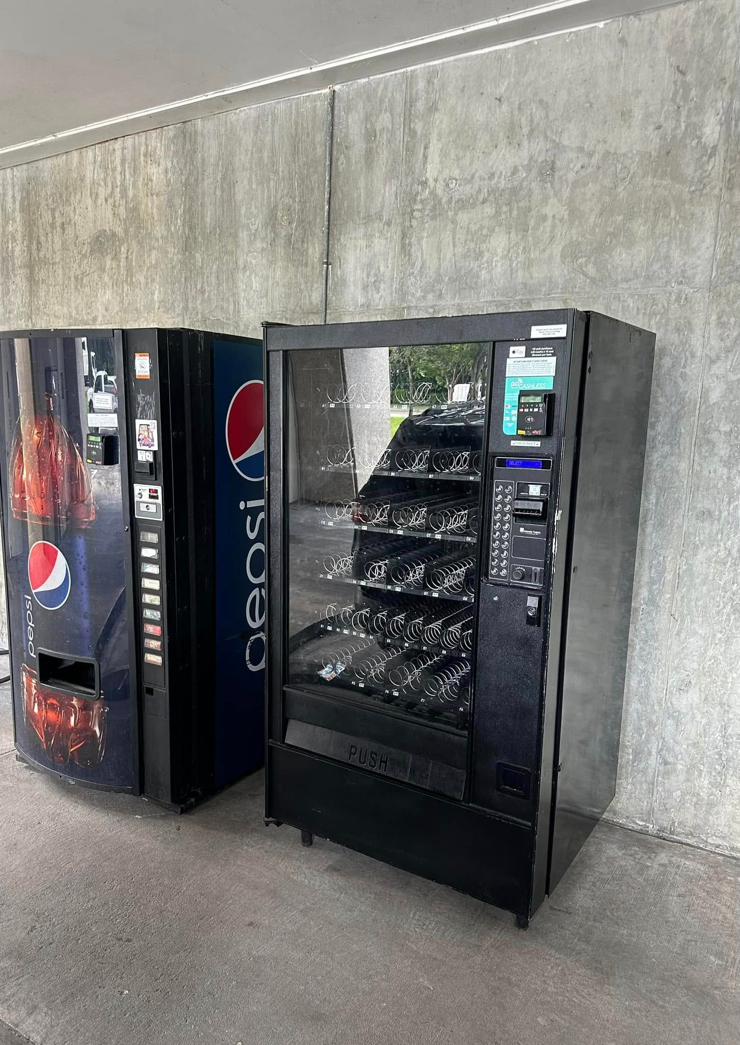 Two vending machines, one branded with a Pepsi logo and a black snack machine, stand against a grey concrete wall.