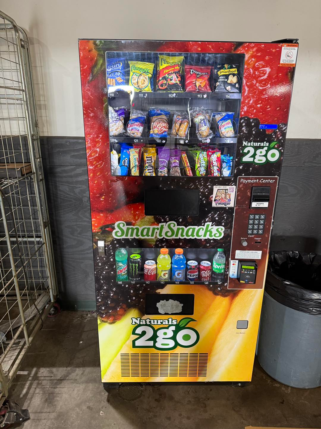 A SmartSnacks vending machine stocked with various chips, snacks, and bottled drinks, standing against a grey wall.