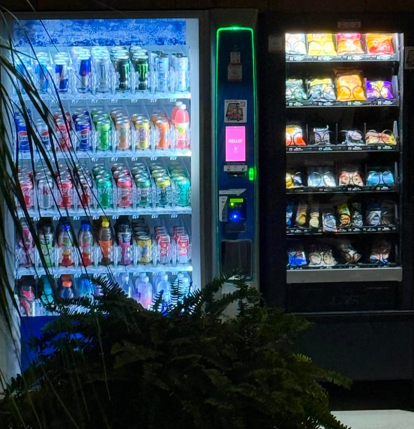 Two vending machines, one filled with various bottled and canned beverages and the other with assorted packaged snacks.