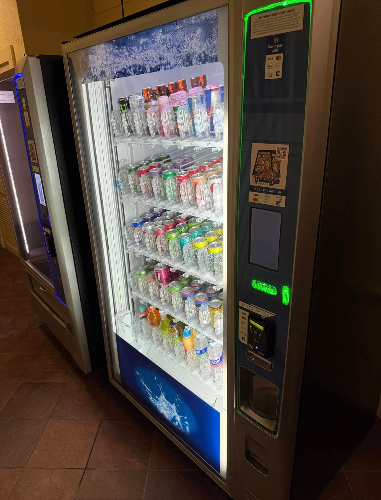 A well-lit, stainless-steel vending machine filled with assorted bottled drinks, positioned indoors on a tiled floor.