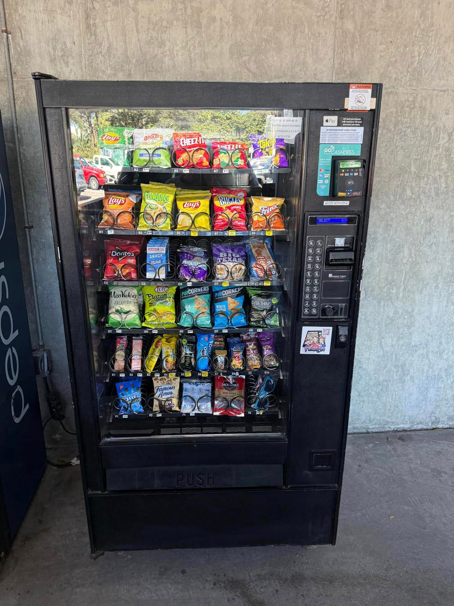A black vending machine stocked with various bags of snacks and chips, standing against a textured wall.