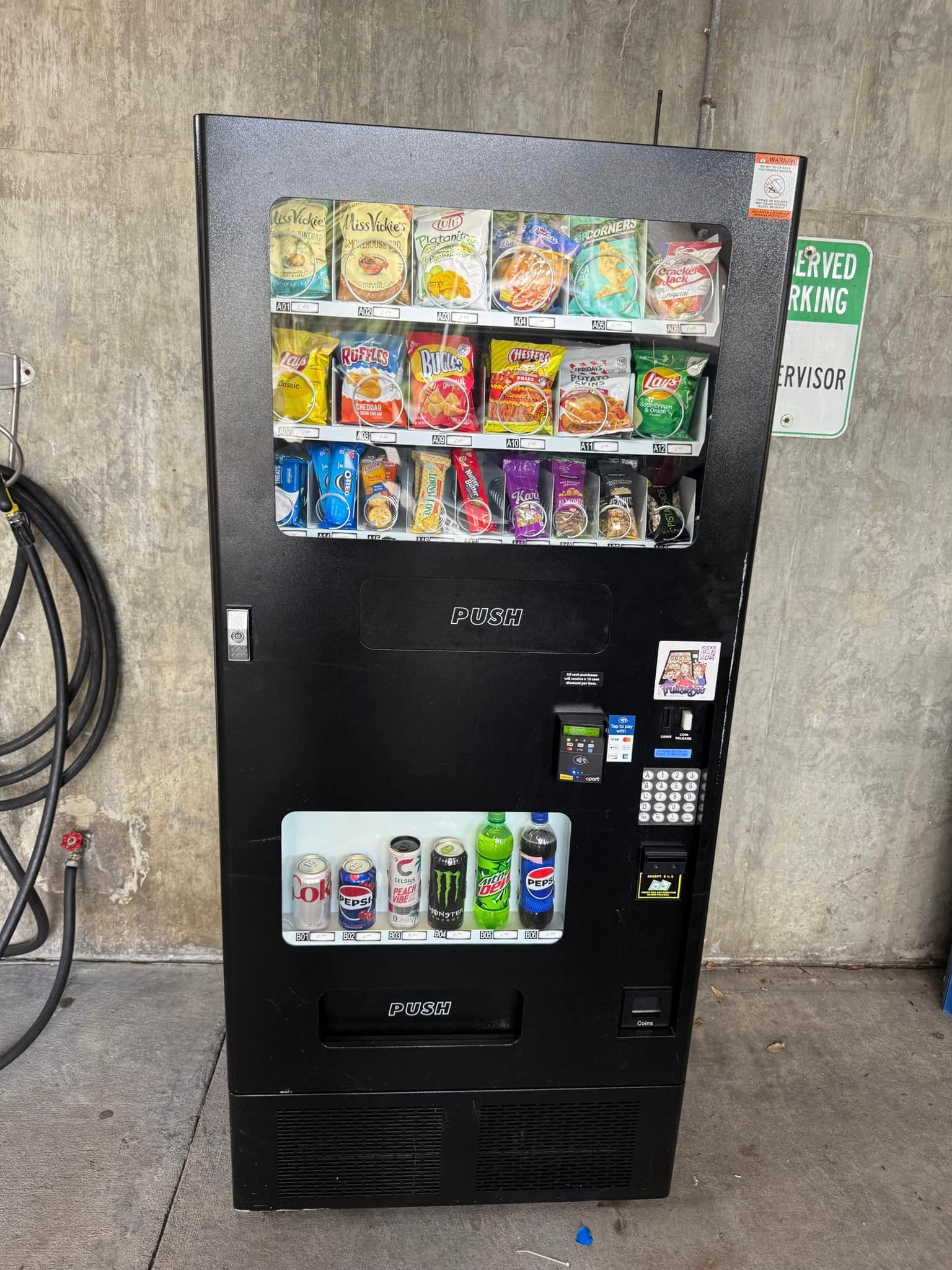 A black vending machine stocked with various snacks and canned drinks, standing against a concrete wall.