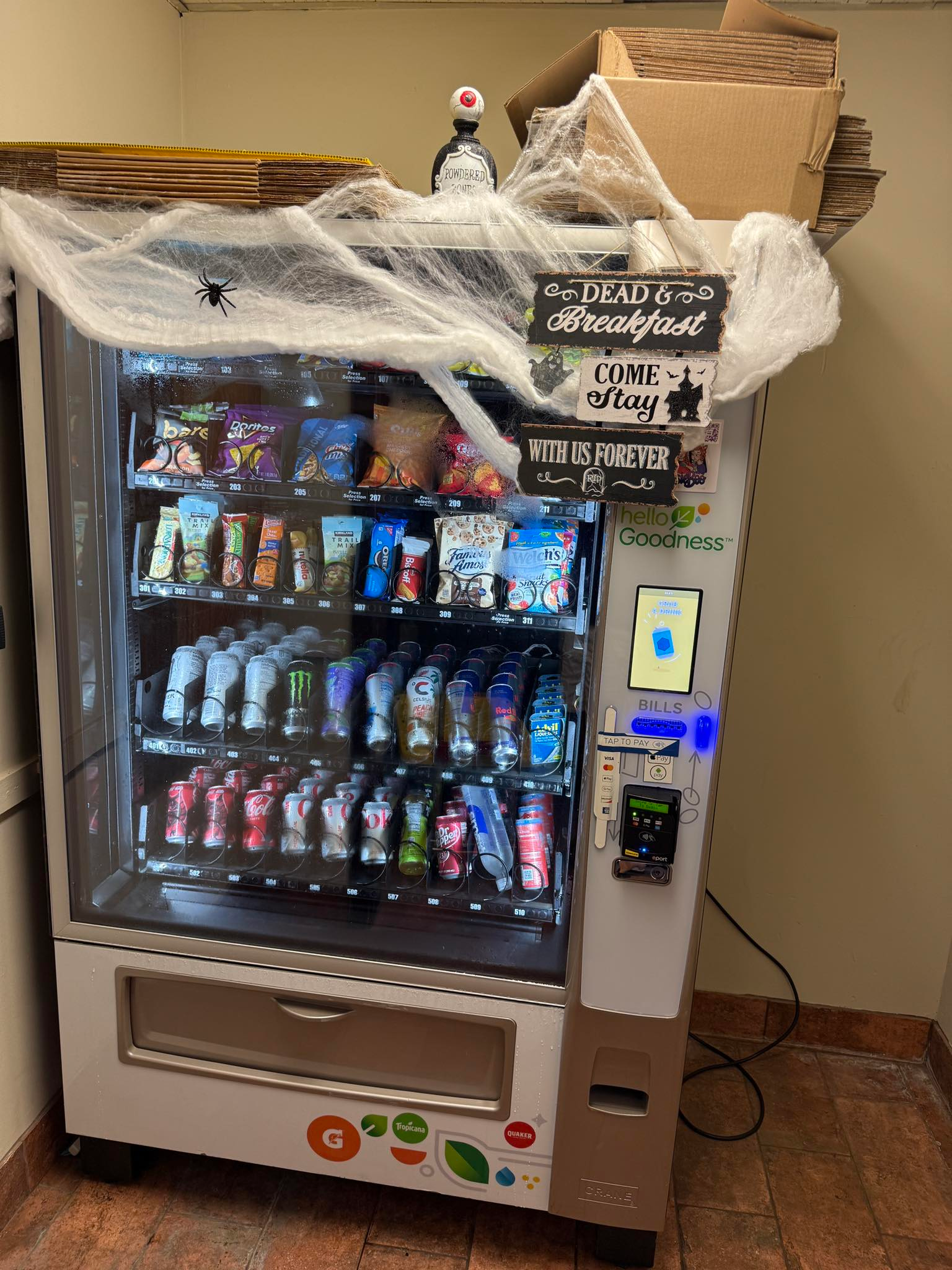 A vending machine decorated with spiderwebs and signs for Halloween, set in an indoor office or breakroom.